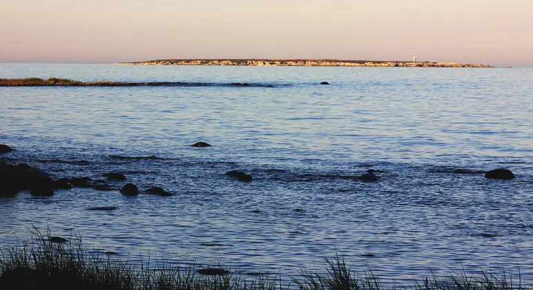 Foto taget från Gotlands kust ut över Östersjön i skymningsljus. Vid horisonten är Heligholmen med sin vita fyr fortfarande solbelyst mot en rosaskiftande himmel.