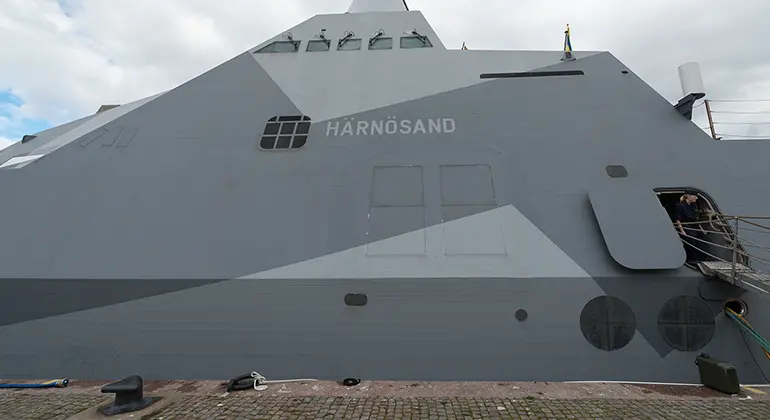 En del av ett modernt militärfartyg. En militär
kliver ut på en landgång till kajen. Fartygets namn ”Härnösand” står på båten.